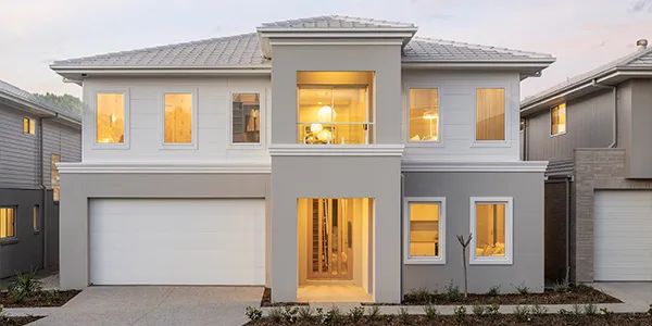 Modern two-story display home with white and gray exterior, featuring a double garage, symmetrical facade with multiple illuminated windows, metal roof, and contemporary landscaping at dusk in Oran Park.