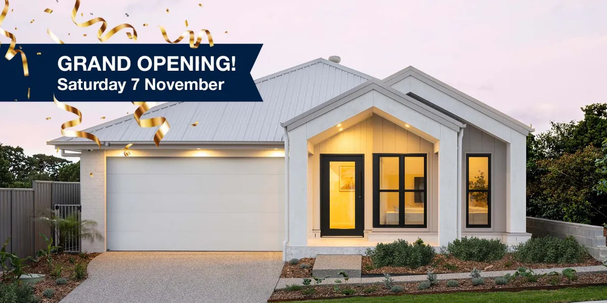 Modern white single-story display home with metal roof, illuminated black-framed windows and yellow door, featuring a grand opening banner with gold confetti graphics reading 