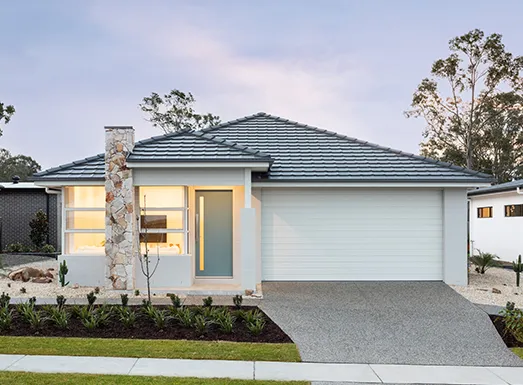 Modern single-story residential home with white rendered walls, dark teal metal roof, and integrated garage. Features large illuminated front windows, a decorative stone chimney accent, and contemporary landscaping with young trees and shrubs along the front garden bed.
