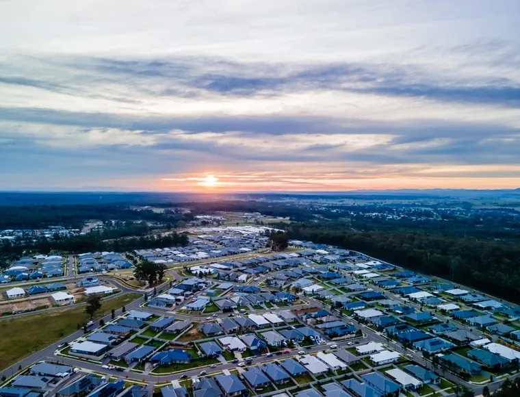 Aerial view of Huntlee Estate residential development at sunset, showing rows of houses with solar panels arranged in a planned community layout, surrounded by forested areas and rolling hills under a dramatic cloudy sky.