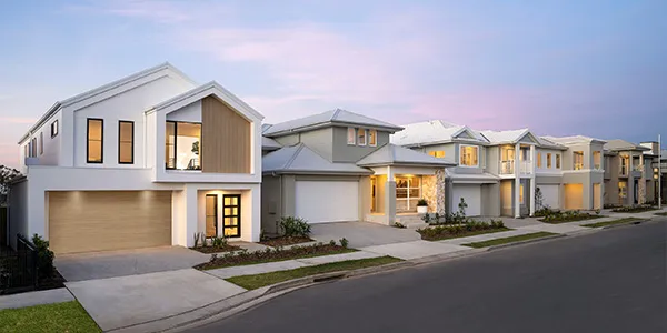 Row of modern display homes in Sydney at dusk, featuring contemporary two-story houses with white and neutral facades, garage doors, illuminated windows, manicured lawns, and a tree-lined street under a pink and purple twilight sky.