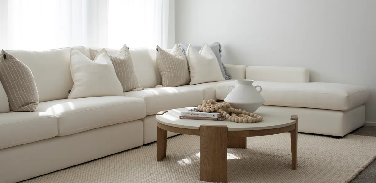Bright, minimalist living room with a cream-colored sectional sofa adorned with neutral and pinstriped pillows, an oval wooden coffee table with a white marble top decorated with books and beaded accessories, and natural light streaming through large windows.