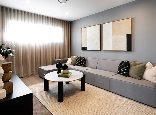 Modern theatre room with gray upholstered seating along the right wall, white round coffee table with black legs in the center, beige area rug, framed abstract artwork on blue-gray walls, floor-to-ceiling beige curtains, potted plant, and minimalist decor in neutral tones.