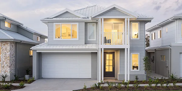 Modern two-story display home in Oran Park with light gray exterior cladding, white trim and columns, pitched roof with metal capping, illuminated windows showing warm interior lighting, white garage door, concrete driveway, and landscaped front garden with young plants.