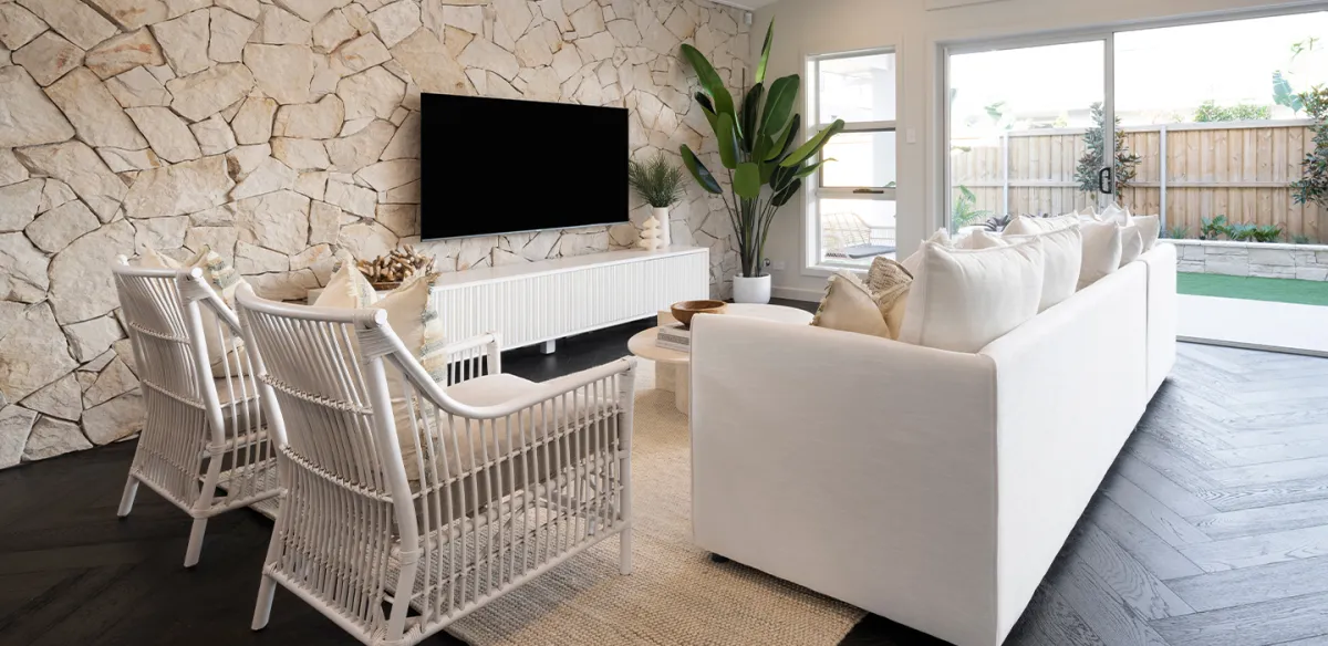 Trinity Leppington display showing a modern living room with cream and white furnishings, including a large sectional sofa and dining chairs, stone feature wall with mounted TV, potted plant, and sliding glass doors opening to a backyard patio.