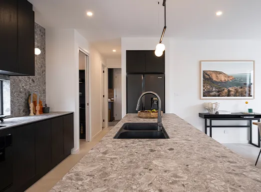 Modern kitchen with black cabinetry, gray speckled countertops, and a black undermount sink with arched faucet. White walls and ceiling with recessed lighting, opening to a hallway and living area with a black console table and coastal landscape artwork.