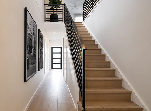 Interior hallway and staircase in a modern home featuring white walls, wooden stairs with black metal railings, light wood flooring, framed artwork on the left wall, and a dark entry door visible in the distance.