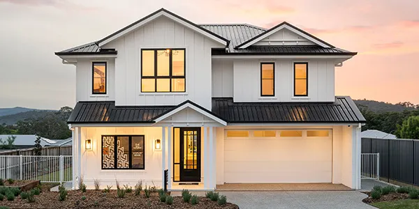 Modern two-story display home with white exterior and dark metal roof, featuring illuminated windows at dusk, garage, front entry with decorative door panel, white metal railing, manicured landscaping, and mountain views in background.