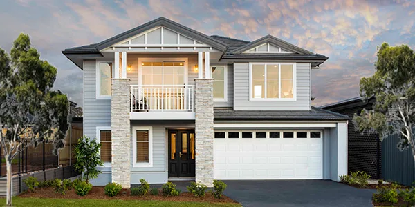 Modern two-story display home with white and light blue exterior, stone accents, dark roof, double garage, front balcony, large windows, and landscaped front yard with trees and garden beds at dusk.