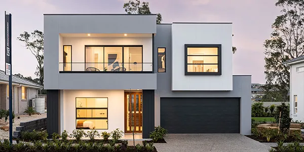 Modern two-story display home with gray and white facade, featuring large windows with warm interior lighting, dark garage door, wooden entry door, and landscaped front garden at dusk.