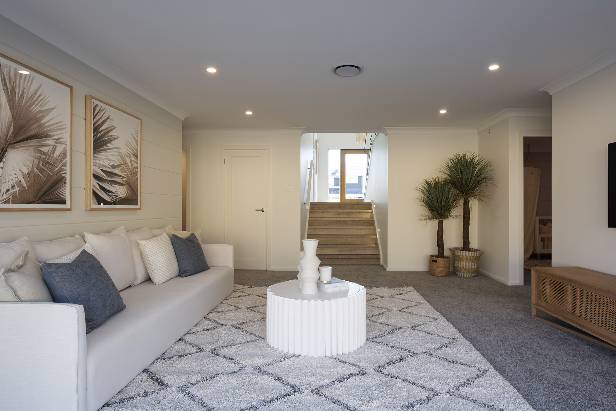 Modern coastal living room with cream-colored sofa featuring blue and white pillows, positioned left side. Center of room displays a white ribbed round table with decorative white objects. Botanical framed artwork hangs above sofa. Wooden staircase with natural wood treads leads upward in background, flanked by potted palm plants in woven baskets. Geometric patterned gray and white area rug anchors seating area. Recessed ceiling lights and