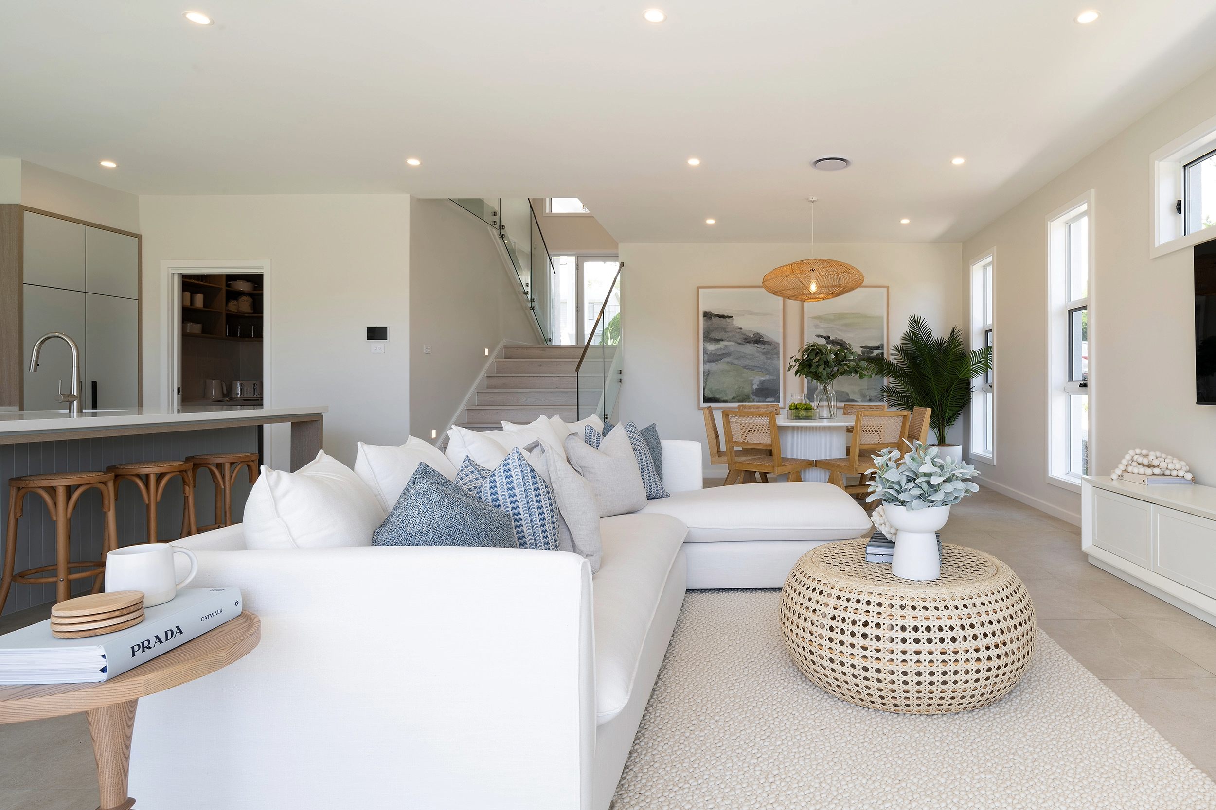 Modern coastal living room with white sectional sofa, blue patterned pillows, and woven pouf ottoman. Open floor plan features a kitchen with wood bar stools on the left, wooden dining chairs and coastal artwork in the background, and a staircase with glass railing. Natural light from multiple windows, recessed ceiling lights, and a woven pendant lamp. Decor includes potted plants, white pottery, and decorative books on wooden side tables.