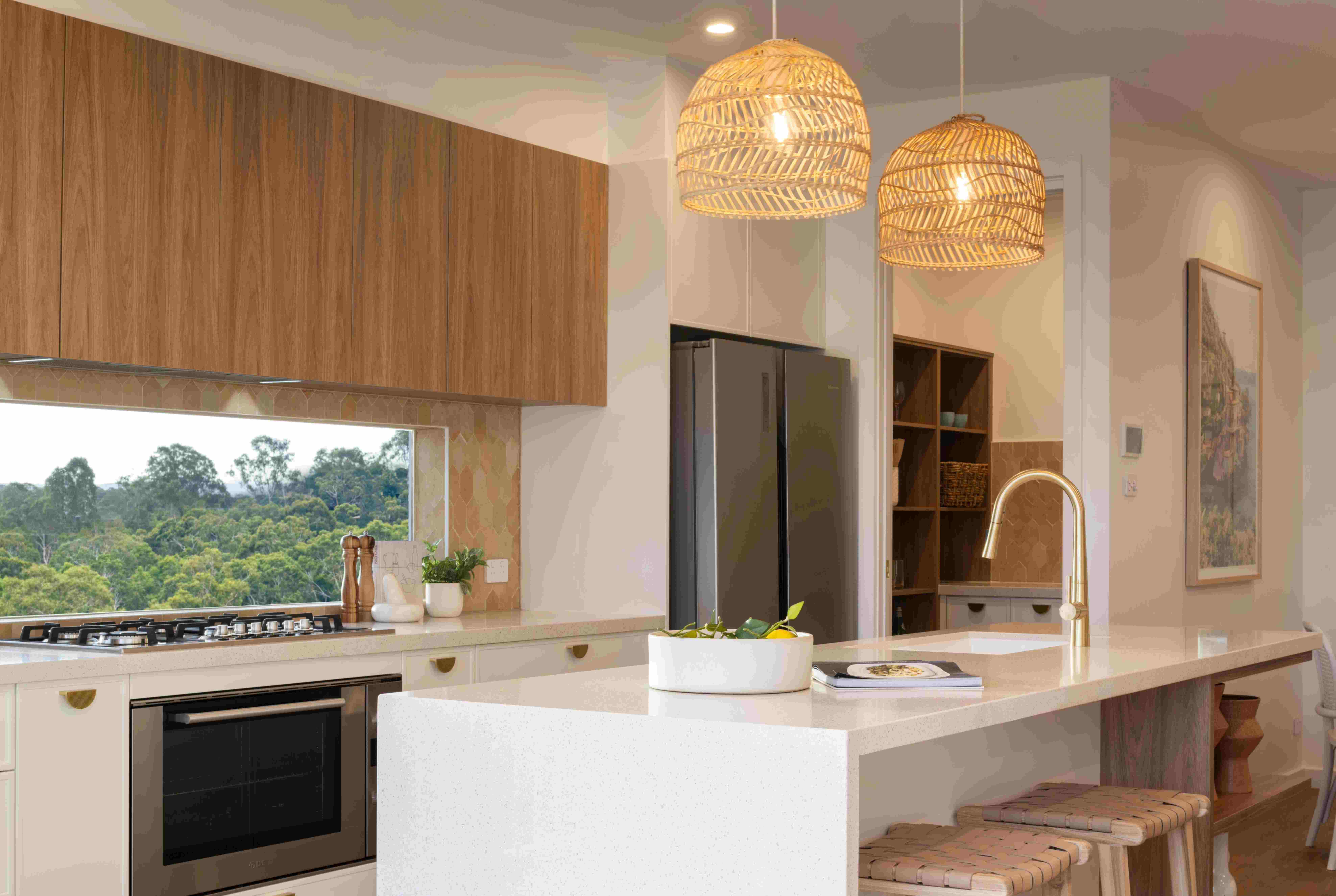 Modern kitchen with white island bench, woven pendant lights, and wood cabinetry. Large window overlooks lush green bushland. Features stainless steel appliances, open shelving, and coastal-inspired design.