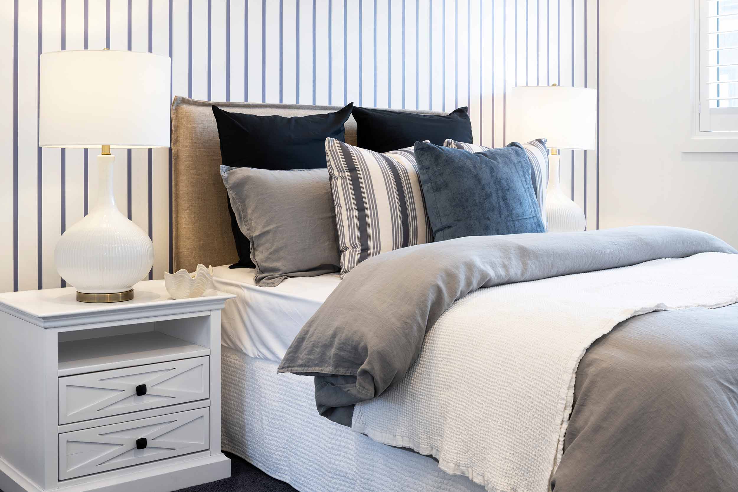 Modern bedroom with a bed featuring layered bedding in neutral tones and black pillows, a striped upholstered headboard, white nightstand with lamp, and vertical striped wallpaper in blue and white tones.