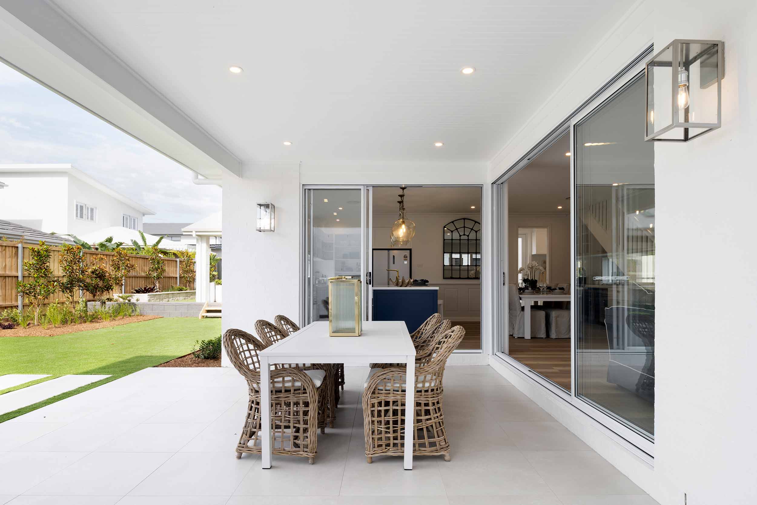 Modern outdoor entertaining area with white dining table and woven chairs on a covered patio, opening via large sliding glass doors to a bright kitchen with blue island, and a manicured backyard with green lawn and wooden fence.