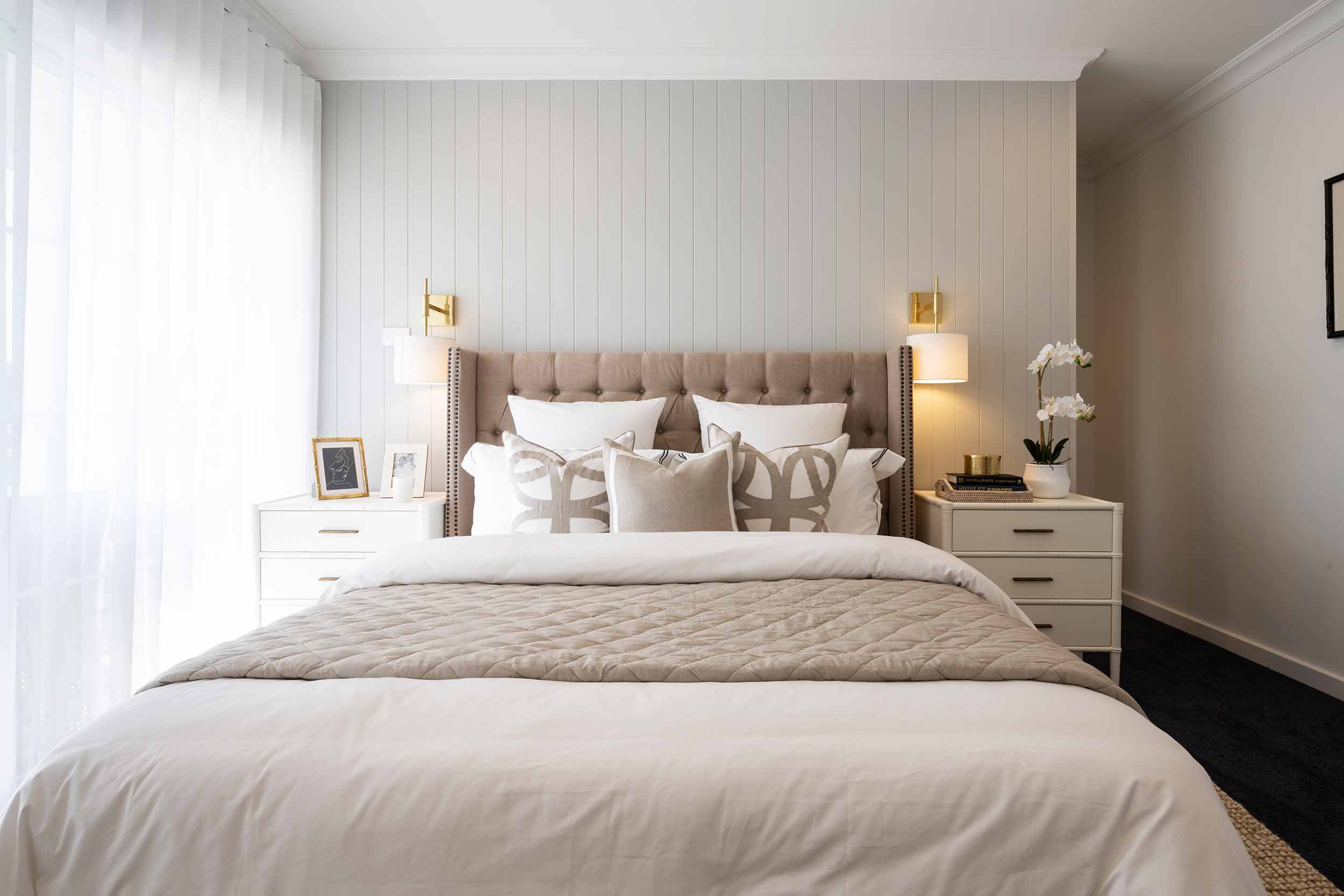 Modern bedroom with a upholstered beige headboard bed dressed in white and neutral bedding, flanked by white nightstands with brass lamps and white flowers. Vertical shiplap accent wall behind bed, large window with sheer curtains on left, and gold wall sconces above nightstands.