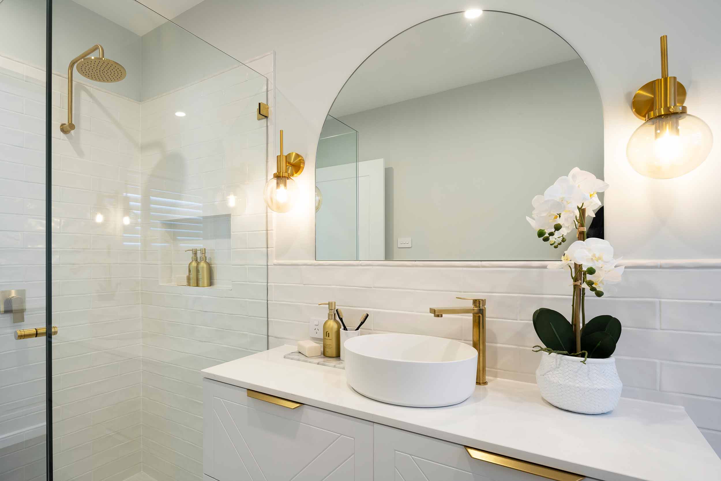 Modern luxury bathroom with white subway tile walls, large arched mirror, and brass fixtures. Vanity features a round white vessel sink, brass faucet, and white orchid plant in decorative pot. Glass-enclosed shower with rainfall showerhead visible on left. Brass pendant lights and recessed lighting illuminate the space. White cabinetry with brass hardware completes the contemporary design.