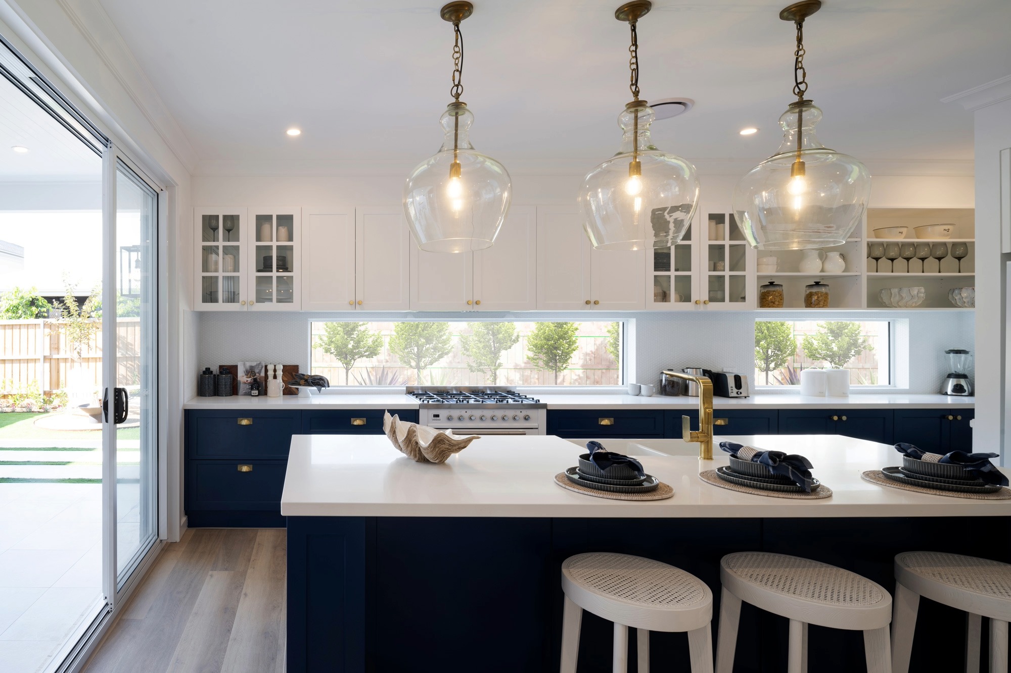 Modern kitchen with two-tone cabinetry featuring white upper cabinets and navy blue lower cabinets with brass hardware. Three clear glass pendant lights hang above a white quartz countertop island with navy blue base. White upholstered bar stools are positioned at the island. The kitchen includes a gas range, white countertops, and windows with garden views. Large sliding glass doors open to an outdoor patio area on the left side.