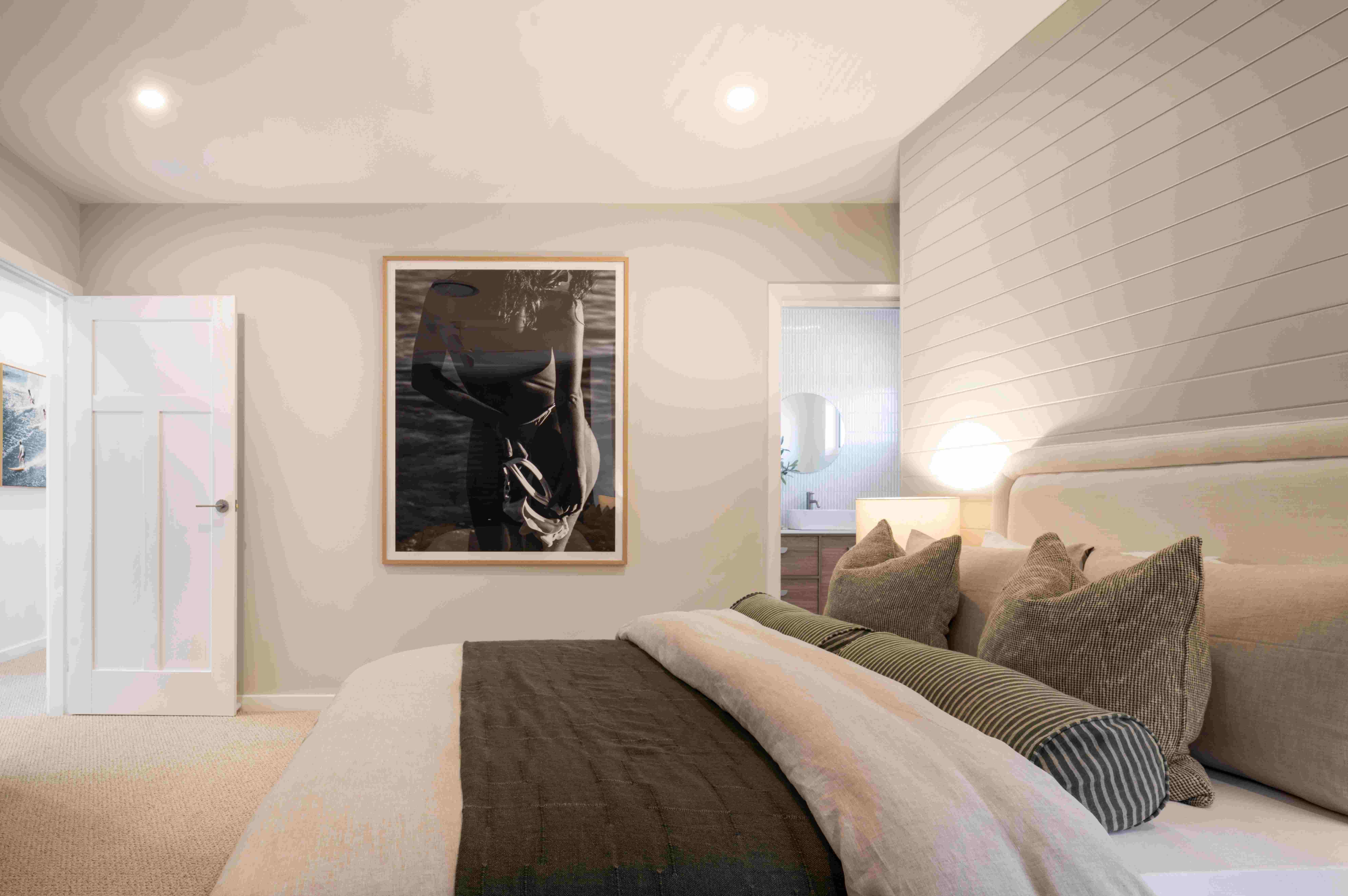 Modern bedroom interior with a bed featuring neutral-toned bedding and decorative pillows, white shiplap accent wall, framed black and white artwork above the bed, recessed ceiling lights, white entry door on the left, and a doorway to an ensuite bathroom on the right.