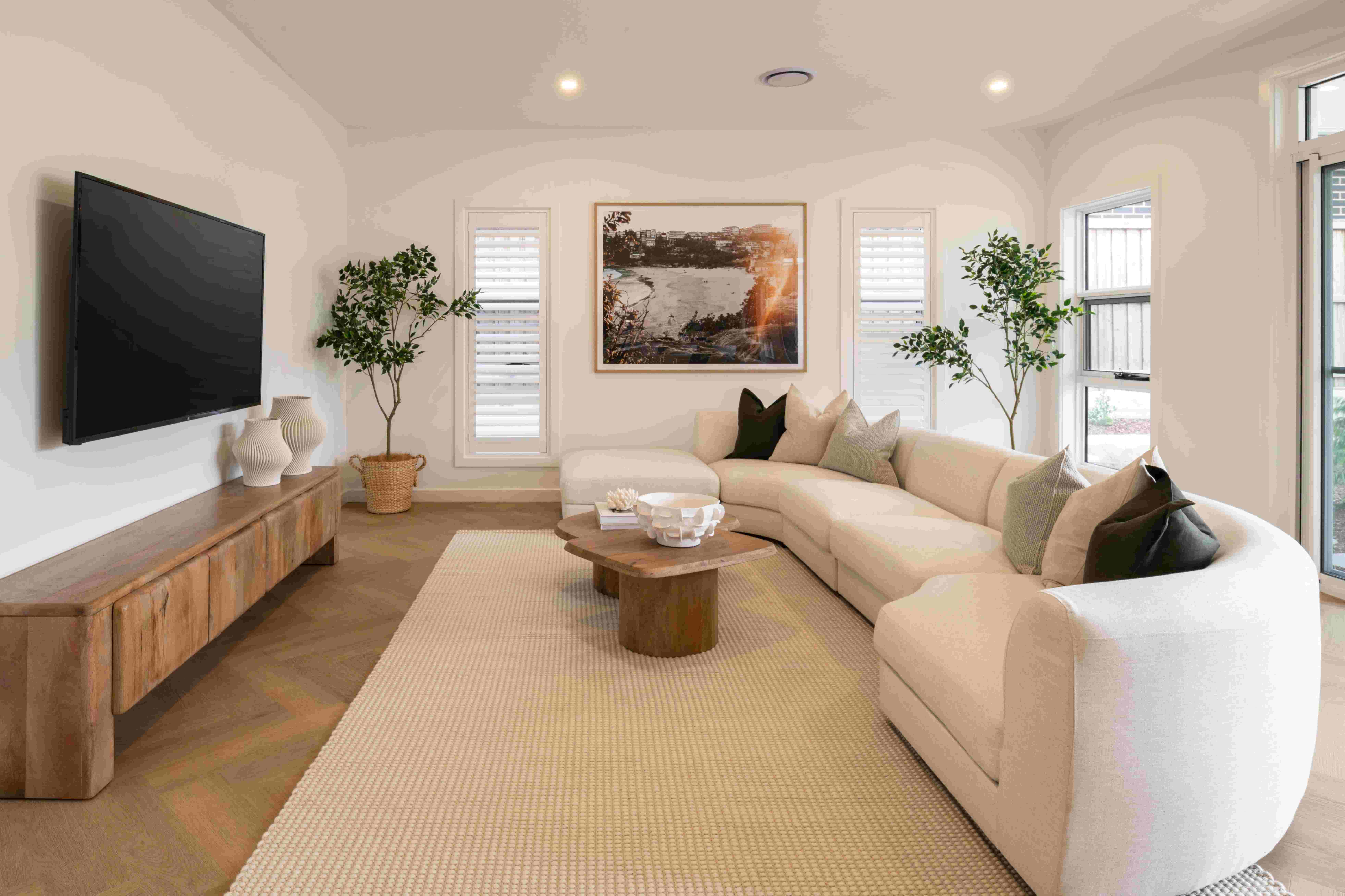Modern living room with curved cream-colored sectional sofa, wooden media console with wall-mounted flat-screen TV, round wood coffee table on neutral area rug, framed landscape artwork, potted plants, white shuttered windows, and recessed lighting in ceiling.
