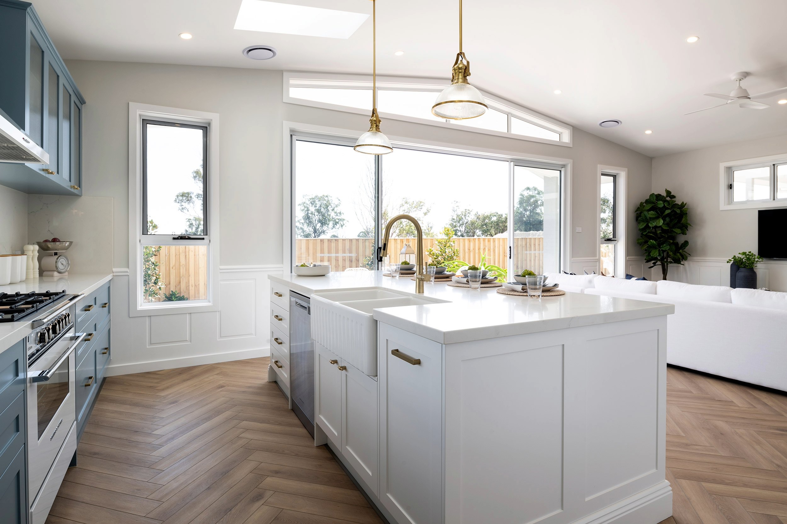 Modern kitchen with white cabinetry and central island featuring a brass sink and faucet, pendant lights overhead, herringbone wood flooring, large windows overlooking a wooden deck, and blue-grey cabinetry on the left with stainless steel appliances. Natural light fills the bright, open-plan space with potted plants and minimal decor visible.