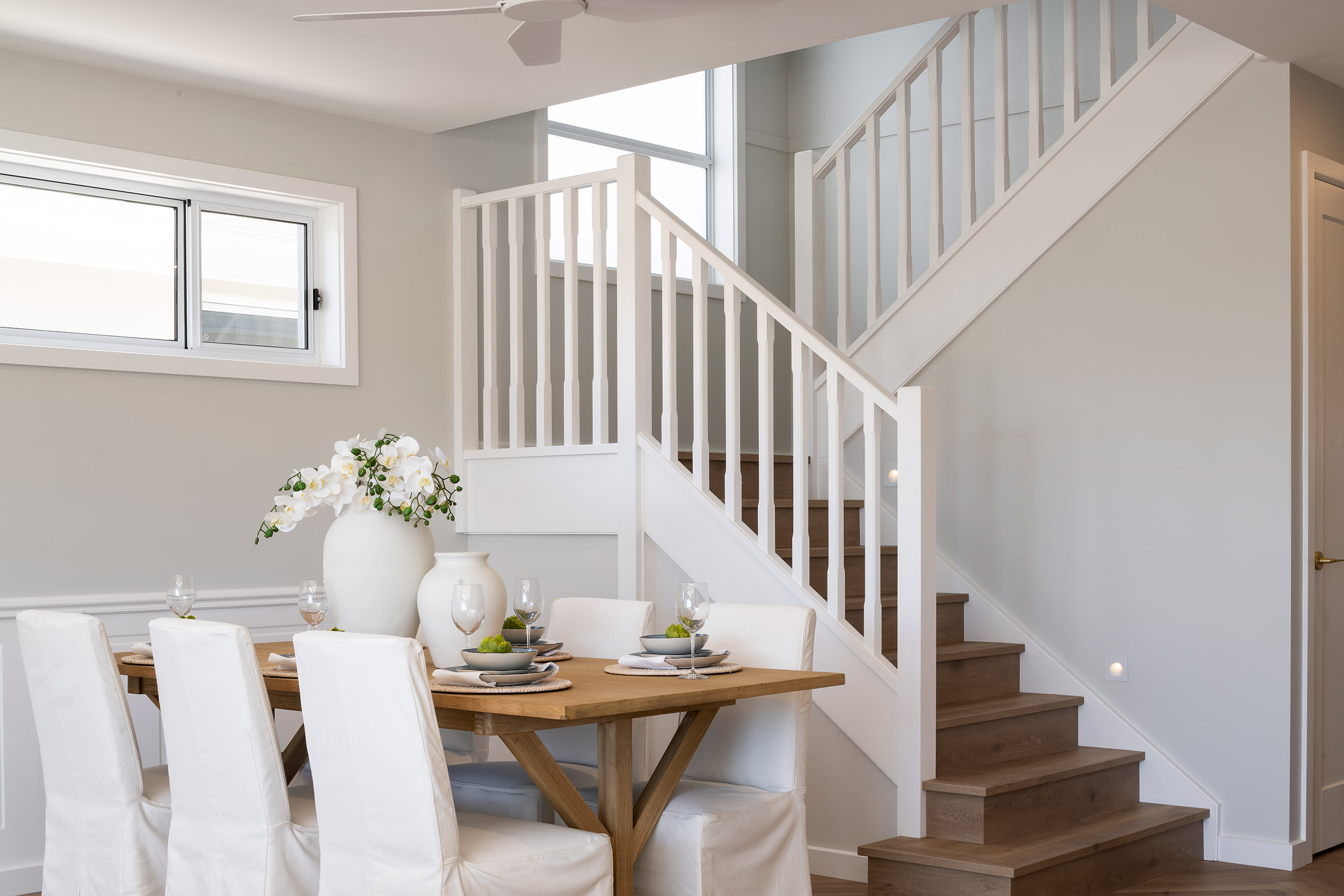 Modern dining area in a two-story home featuring a wooden table with white upholstered chairs, white flower arrangement centerpiece, and a staircase with white railings and wooden steps leading to the upper level. Large windows provide natural light to the neutral, minimalist interior.