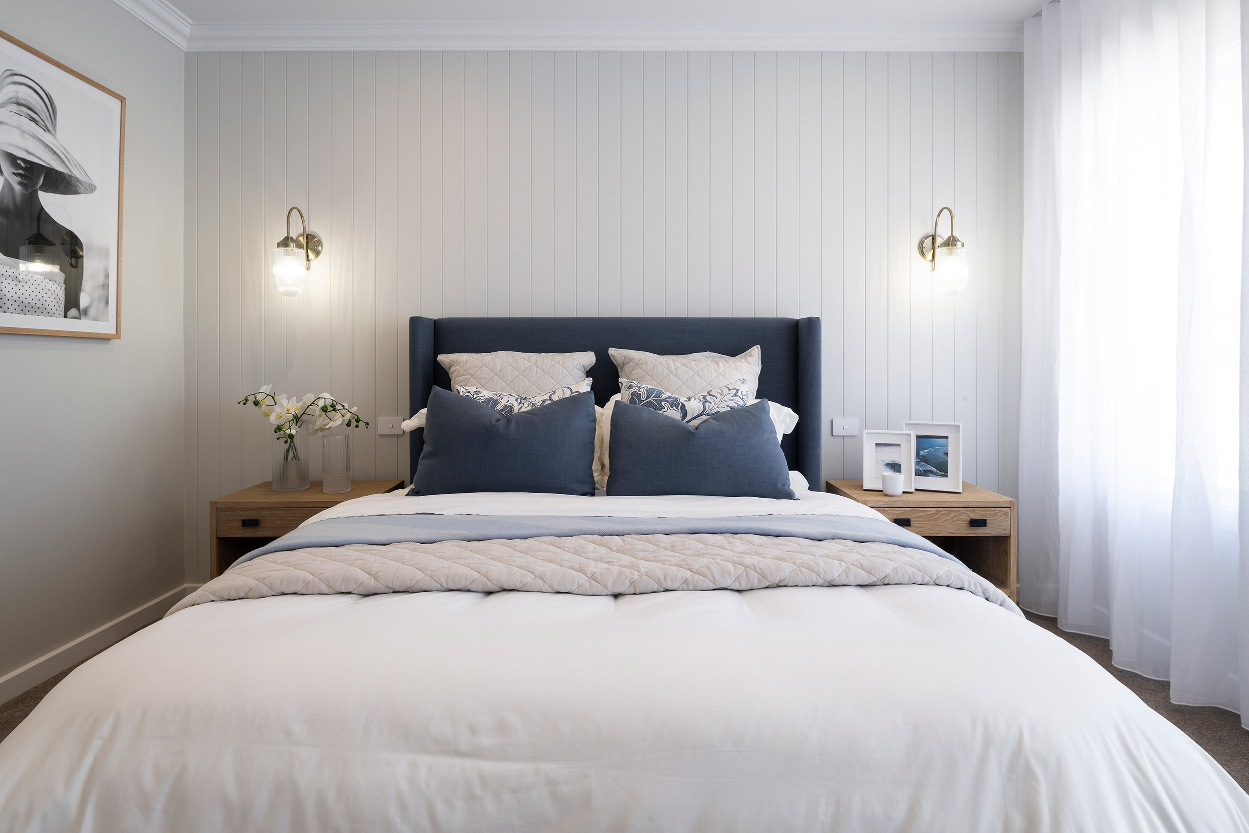 Modern bedroom with a dark navy upholstered bed featuring white and patterned pillows, centered against a white vertical shiplap wall. Warm brass pendant lights flank the bed. Wood nightstands with fresh flowers and decor sit on either side. White bedding with a quilted throw covers the bed. A framed black and white portrait hangs on the left wall, and sheer white curtains frame a window on the right. Minimalist, contemporary design