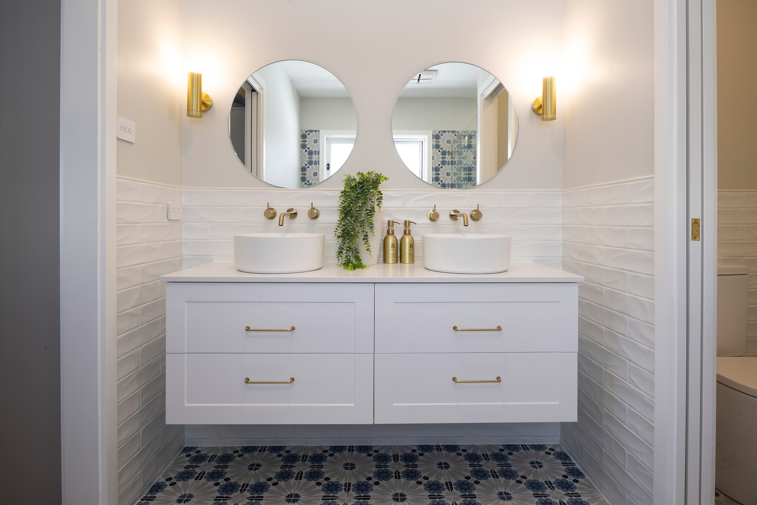 Modern bathroom vanity with dual round mirrors, two white vessel sinks, white cabinetry with brass hardware, brass wall sconces, white subway tile backsplash, brass faucets, potted green plant, and patterned floor tile in blue and white geometric design.