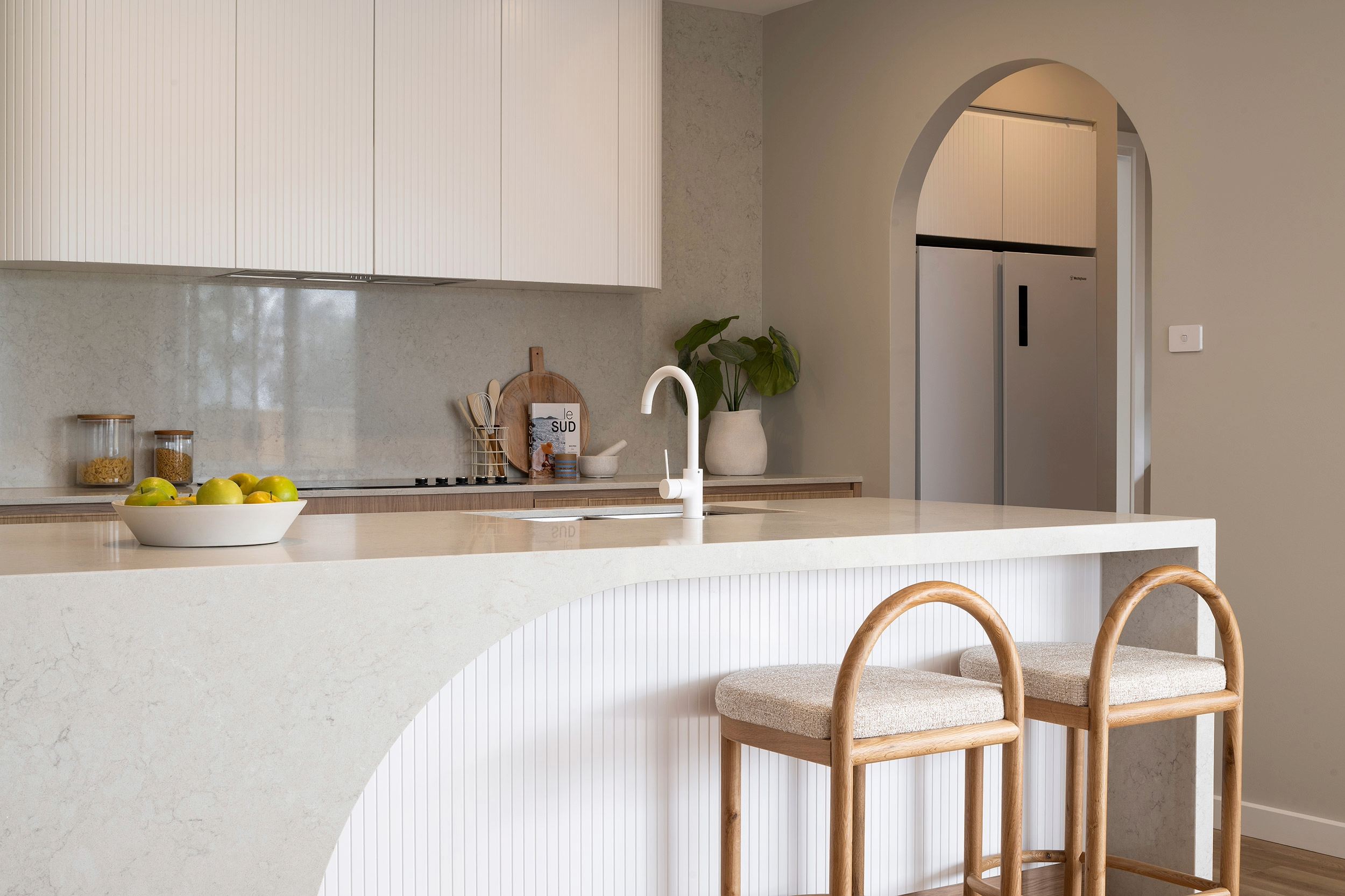 Modern luxury kitchen with curved white marble island featuring two wooden bar stools with beige cushions. White cabinetry, white faucet, and potted plant on countertop. Arched doorway visible in background. Bowl of lemons on counter. Minimalist design with neutral tones.