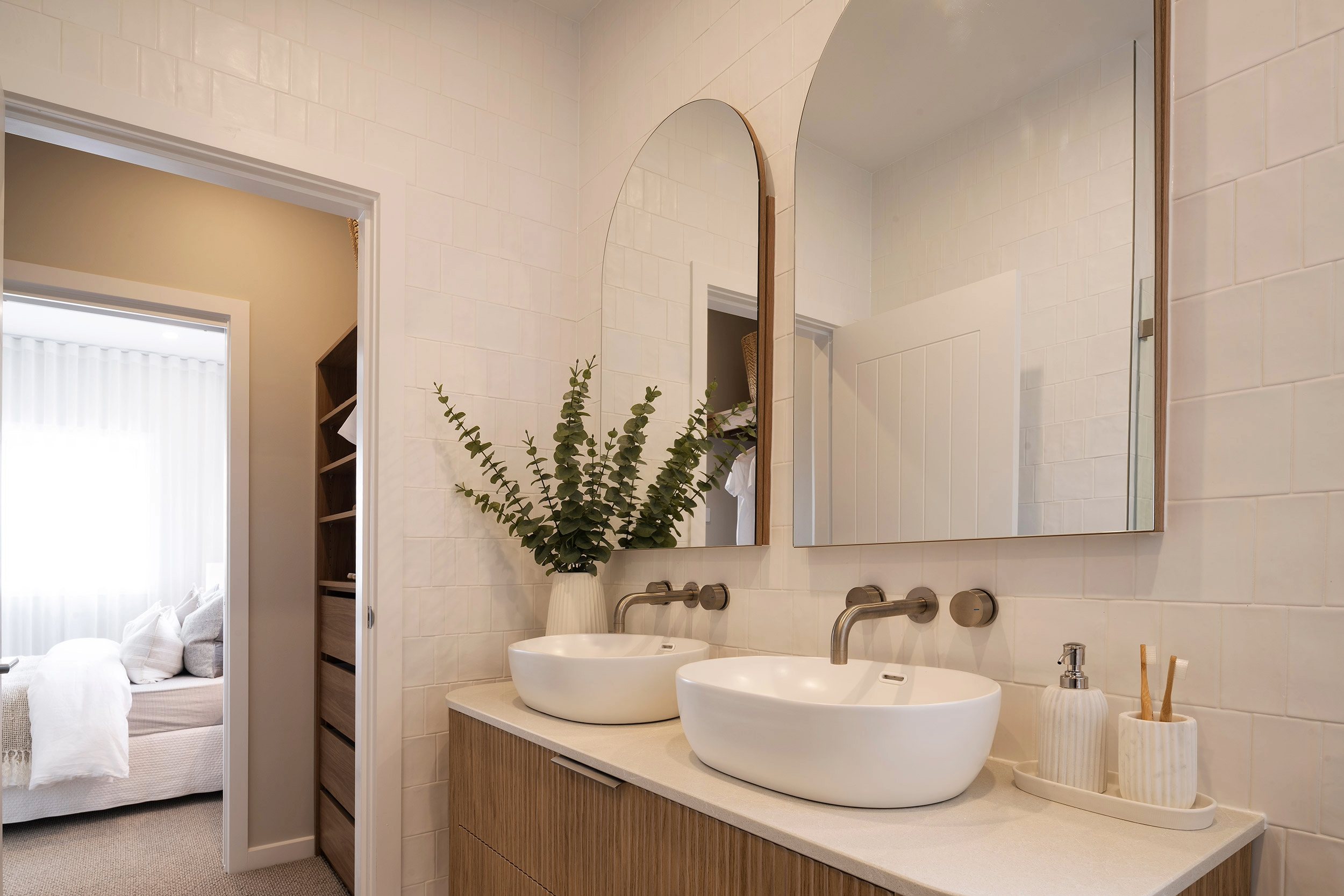 Luxury bathroom with dual white vessel sinks on a light wood vanity, arched and rectangular mirrors above, brushed bronze fixtures, cream subway tile walls, fresh eucalyptus arrangement in a vase, and a view into an adjoining bedroom with white bedding.