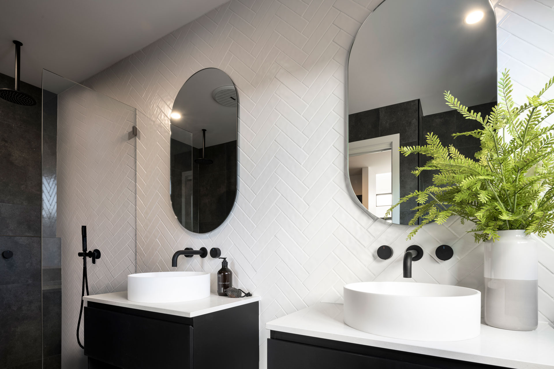 Modern luxury bathroom with white herringbone tile walls, two white cylindrical vessel sinks on black vanities with matte black fixtures, oval and round mirrors, and a potted fern plant. Dark shower enclosure visible on the left with black rainfall showerhead.