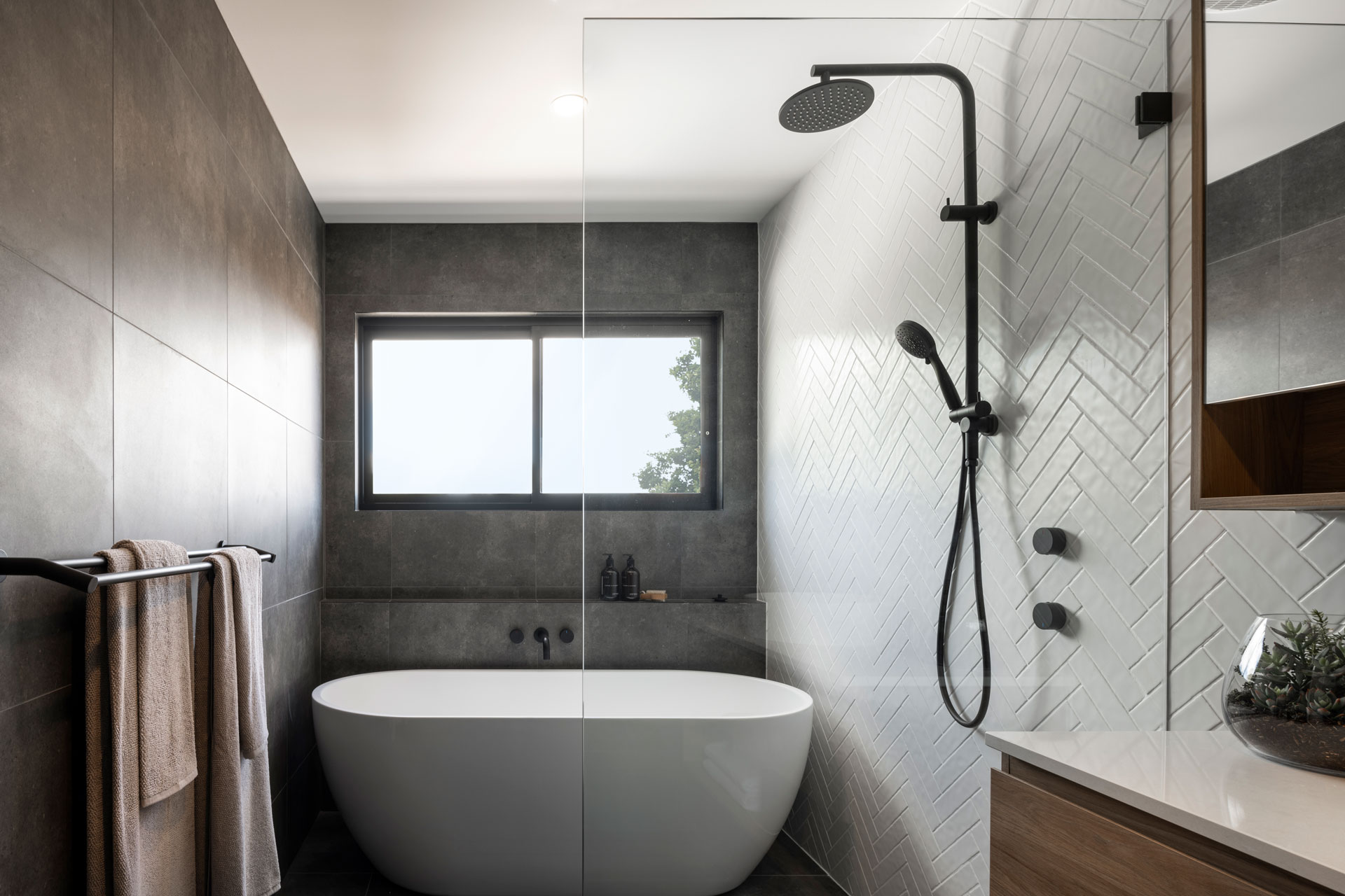 Modern bathroom with a freestanding white oval bathtub centered in the room, flanked by a walk-in shower with herringbone tile wall and black fixtures on the right, and a towel rack with folded towels on the left. A large window with dark frame overlooks trees, set into a dark gray tile accent wall behind the tub. White subway tiles cover the shower surround, with a wood-look vanity visible on the right edge and a potted