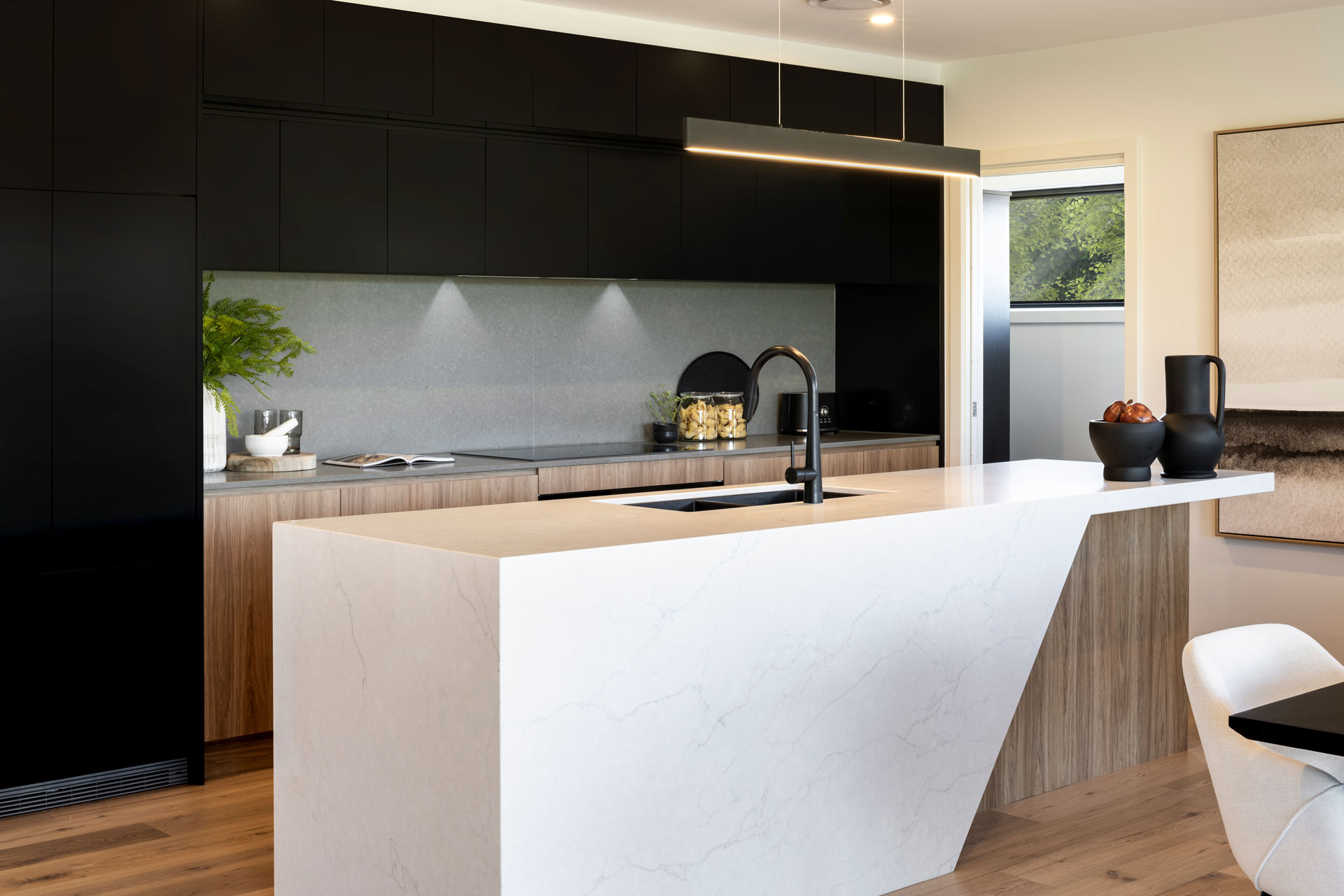 Modern kitchen with white marble island countertop in foreground, black matte upper cabinets with integrated lighting, light wood lower cabinetry, gray tile backsplash, black arc faucet, and stainless steel sink. Wood flooring, cream walls, and natural light from window visible in background. Decorative black vessels and potted plant on counter.