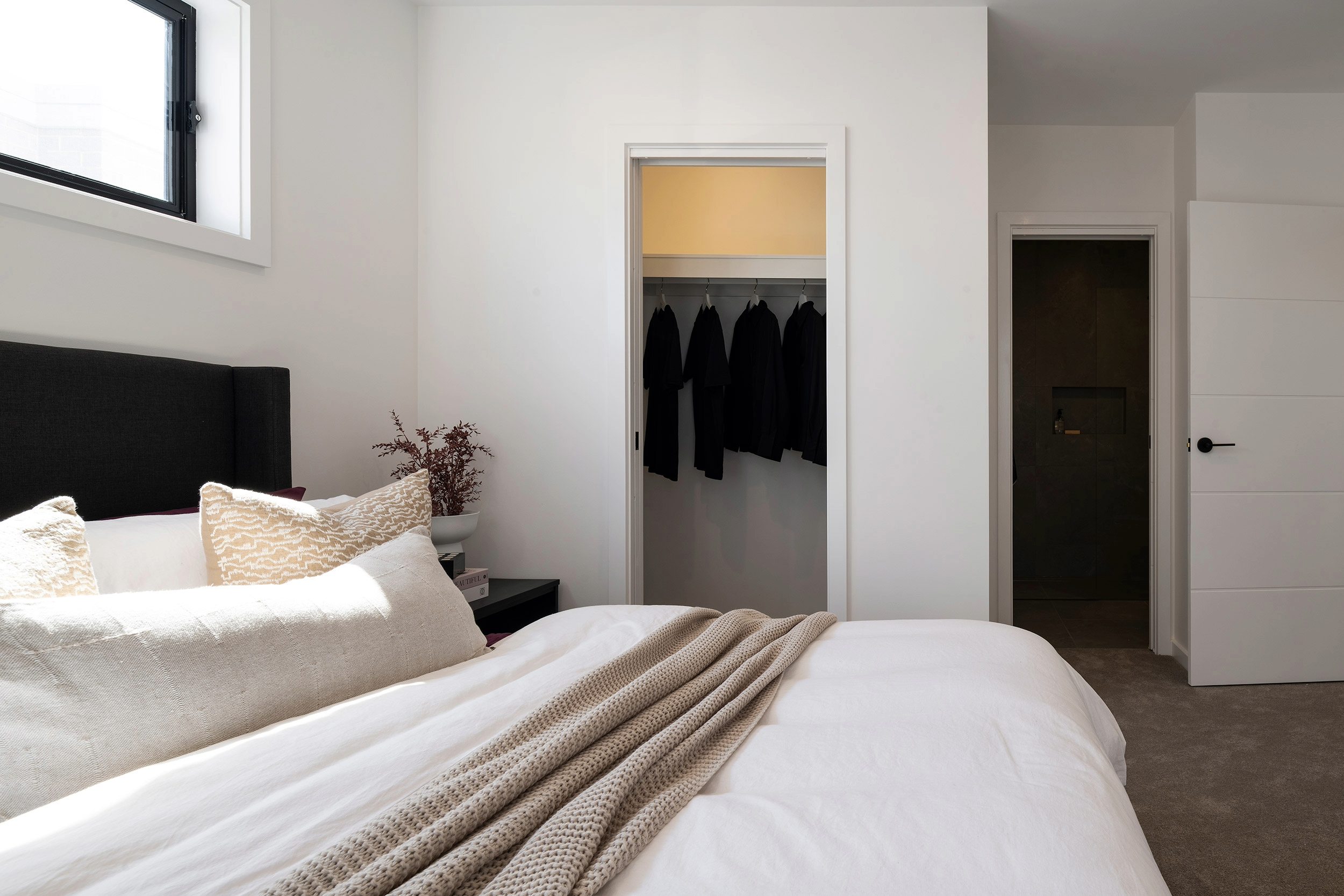 Modern bedroom with queen bed featuring white linens and neutral bedding, black upholstered headboard, and gold patterned pillow. Open walk-in wardrobe with warm lighting displays hanging black clothing. White walls, high window with black frame on left, dark wooden door on right, and potted dried plant on nightstand.