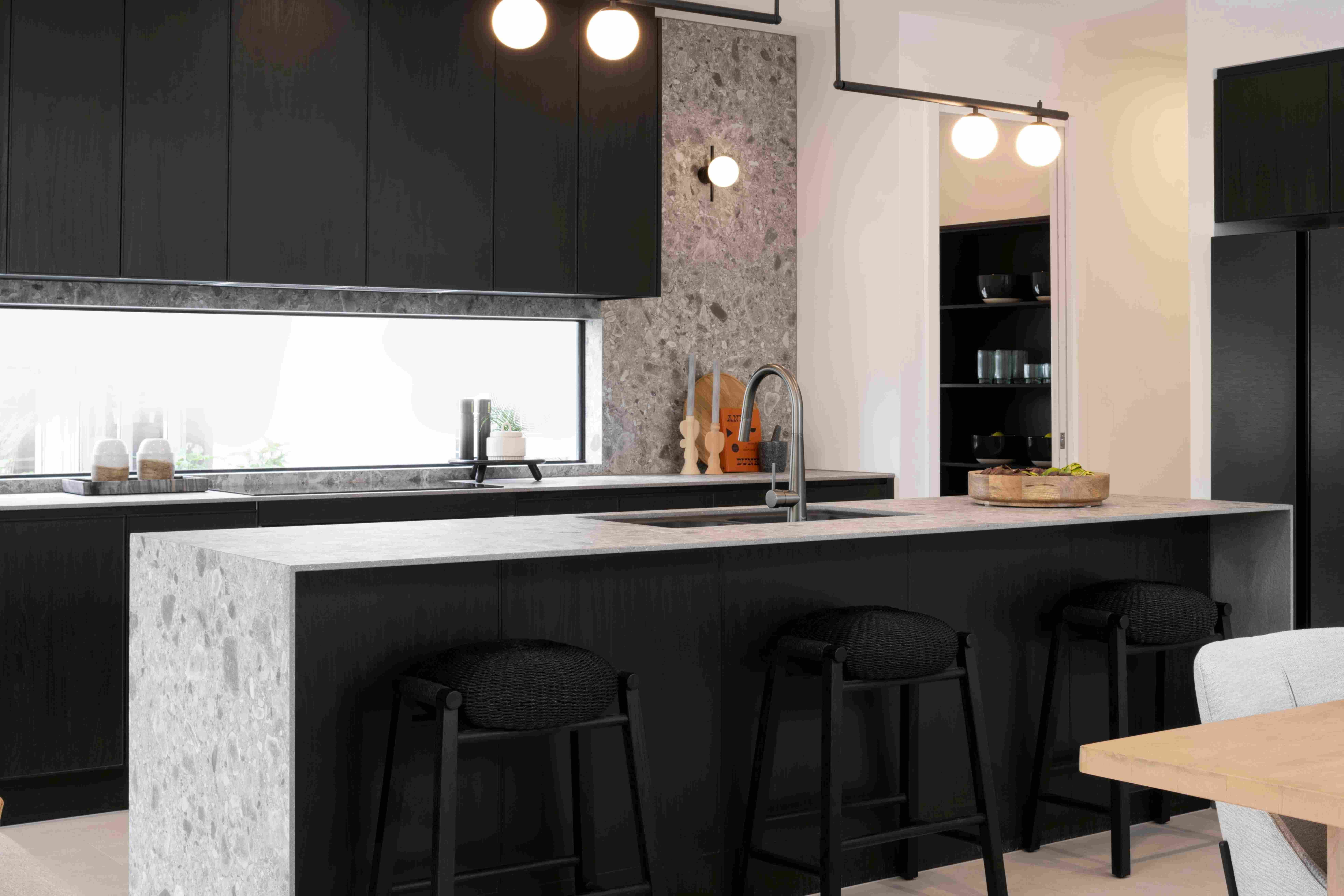 Modern kitchen with black cabinetry, white marble countertops, and a large island with black bar stools. Features a window above the sink with city views, pendant lighting, gray stone accent wall, open shelving, and minimalist design aesthetic.