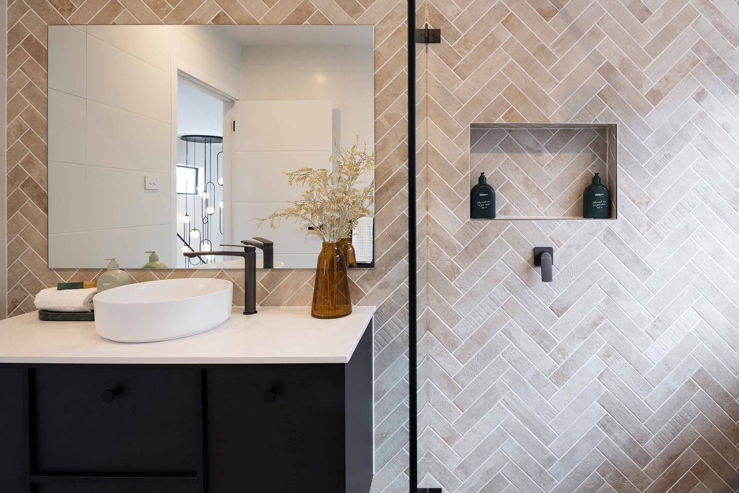 Modern bathroom with herringbone tile walls in taupe and white, featuring a white oval vessel sink on a black vanity with white countertop, large framed mirror, black faucet, amber glass vase with dried flowers, recessed shower niche with dark bottles, and black shower frame visible on the right.
