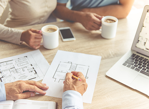 Two people reviewing architectural floor plans and blueprints at a wooden table with coffee cups, a smartphone, and a laptop during a design or construction meeting.
