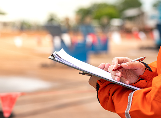 A surveyor in an orange safety vest holds survey papers and a pencil while reviewing contour survey data on a construction or land survey site, with surveying equipment and markers visible in the blurred background.