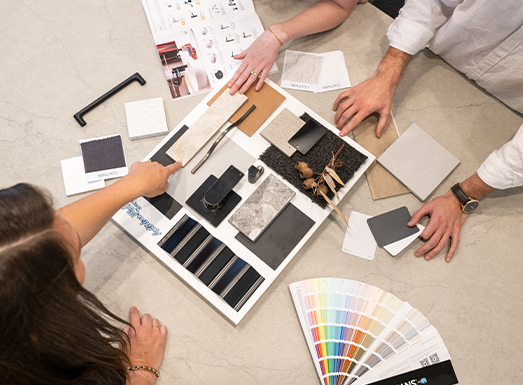 Two people reviewing interior design material selections laid out on a concrete surface, including fabric swatches, color palette fan, wood samples, and black and white material samples arranged in an organized grid layout.
