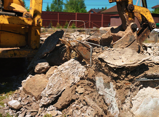 Yellow demolition excavator with claw attachment processing concrete and debris at a demolition site, with crushed rubble and broken concrete pieces scattered across the ground under clear blue sky.
