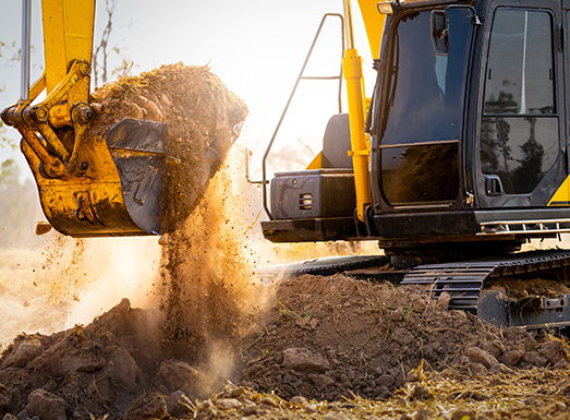 Yellow excavator dumping soil from its bucket during site preparation work, with dust billowing in the air and the machine's cab visible on the right side of the image.