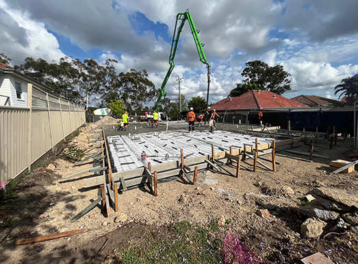 Construction site with concrete foundation being poured using a green pump truck with extended boom. Workers in high-visibility vests oversee the operation on a dirt lot surrounded by metal fencing, existing buildings, and trees under partly cloudy sky.