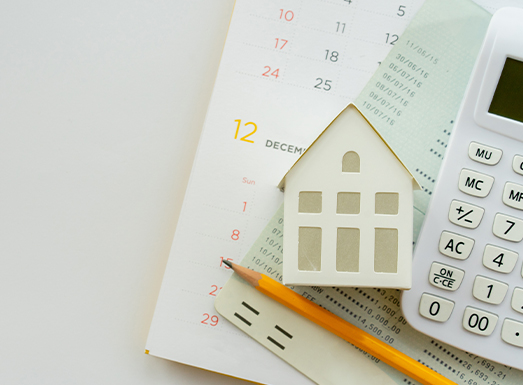 A wooden house-shaped cutout placed on an open calendar showing December dates, alongside a yellow pencil and a white calculator on the right side, arranged on a light background.