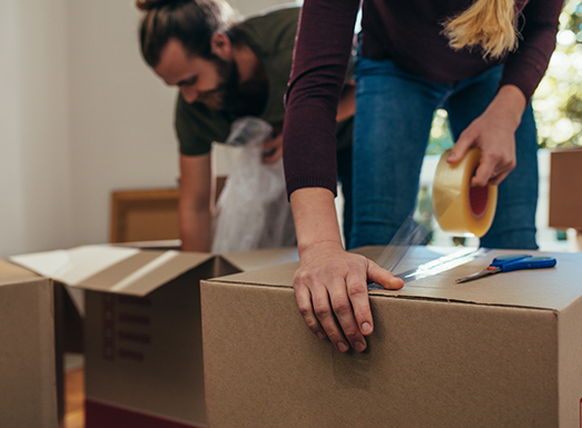 Two people unpacking cardboard moving boxes in an empty room, with one person's hand resting on a box in the foreground.