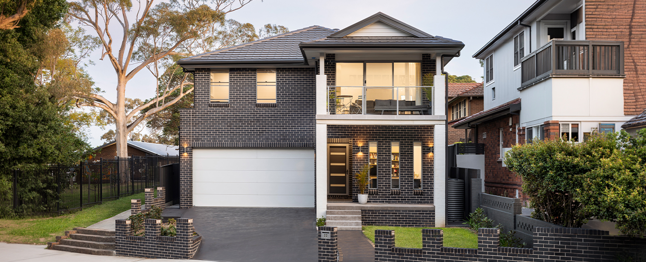 Modern two-story brick house with dark charcoal facade, white trim, and large windows. Features a white garage door, timber front door, and landscaped front yard with native tree and hedging. Suburban street setting with neighboring homes visible.