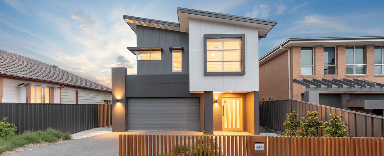 Modern two-story house in Adamstown with contemporary design featuring white upper level and dark gray lower level, illuminated entry door, garage, metal fencing, and neighboring brick homes visible on either side at dusk.