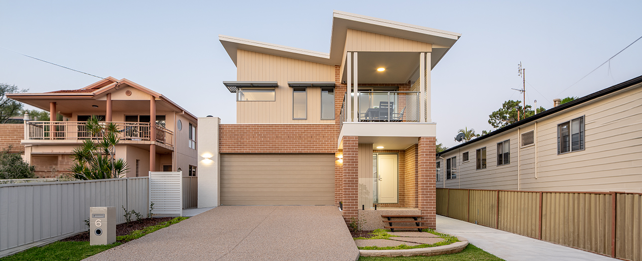 Modern two-story brick house with contemporary architecture, featuring a garage, illuminated upper-level balcony, and driveway. Neighboring older residential homes visible on either side. Clear evening sky with outdoor lighting highlighting the entrance.