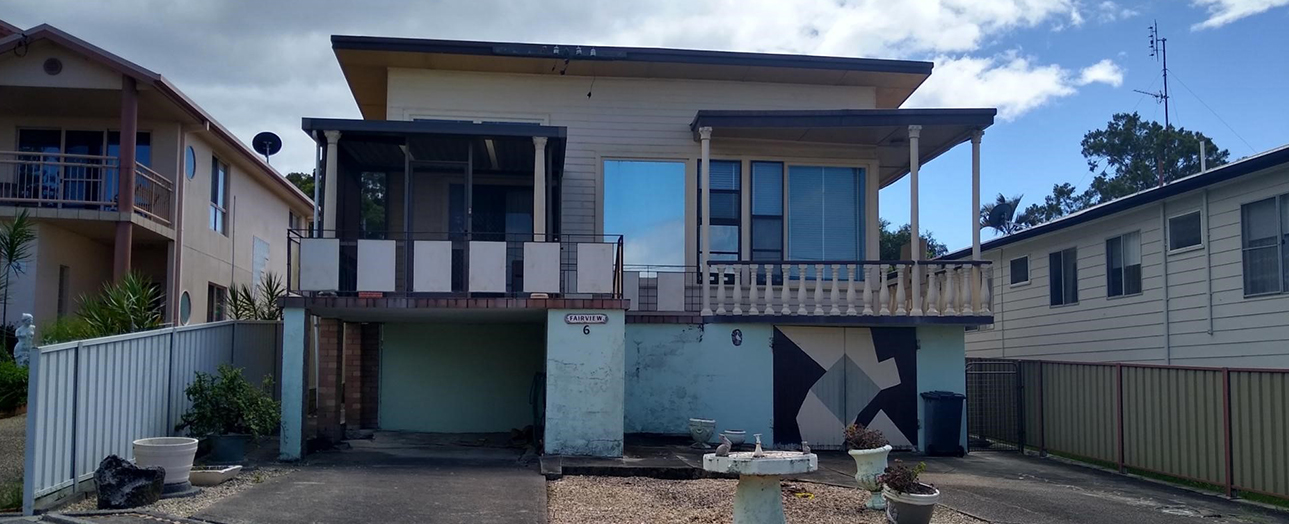 Weathered elevated house with blue and white exterior, showing signs of age and wear. The single-story dwelling features a covered front porch, multiple windows, and metal railings. The foundation appears elevated on stilts with deteriorating blue-painted underside panels. Neighboring houses visible on either side. Daytime photograph with partly cloudy sky.