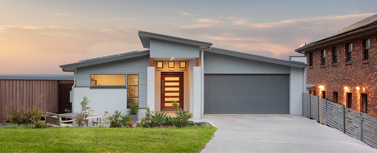 Modern single-story white house with gray roof and wood grain garage door at dusk, featuring illuminated front entrance, landscaped garden with potted plants, concrete driveway, and adjacent brick residence on the right.
