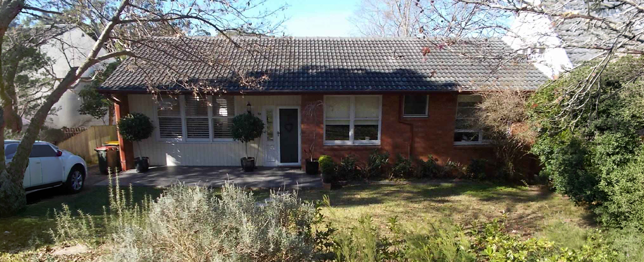 Single-story brick residential house with tiled roof and attached garage, featuring an overgrown front yard with wild grasses and sparse vegetation, bare trees overhead, and a white car parked on the left side of the driveway.
